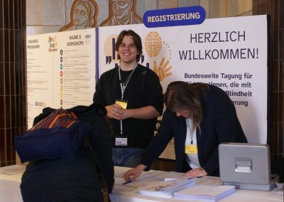 Blick auf den Registrierungstisch für die Tagung. Dahinter steht ein Banner, welches die Teilnehmer*innen willkommen heißt. Zwei Mitarbeiter des BBI sind fleißig bei der Arbeit.