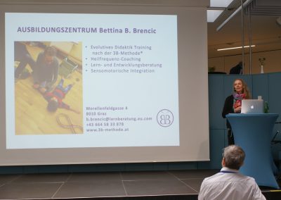 Frau Brencic hält den dritten Vortrag des Tages. Sie steht am Rednerpult, daneben wird ihre Präsentation gezeigt. Tagungsleiter Hr. Weingartner ist im Vordergrund im Publikum zu sehen.
