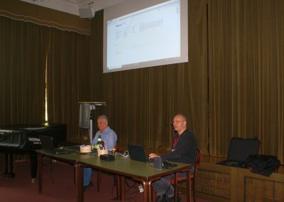 Workshop 8 von Mag. Schmid und Herrn Vetter, MA. Beide sitzen im Festsaal auf einem Tisch mit Laptops und Lautsprechern unter einer Präsentations-Leinwand. Sie sind bereit zu Musizieren!